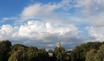 Blauer Himmel mit Wolken, unten grüne Bäume und Kirchturm